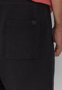 Schwarze Shorts mit einer Gesäßtasche und einem eingenähten Etikett. Der Stoff hat eine glatte Textur und einen elastischen Bund. Das Gesamtdesign ist schlicht.
