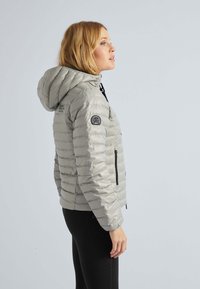 Lys grå pufferjakke med hætte, med lodret quiltet design, frontlynlåse og et sort logo-patch på ærmet.