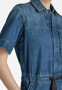 Une combinaison en denim de lavage bleu moyen, avec un col, des manches courtes, des poches poitrine et une taille nouée pour souligner la silhouette.