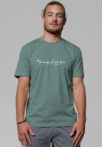 Groene katoen t-shirt met witte tekst die "So einfach ist das" op de borst leest. Korte mouwen en een klassiek rond decolleté ontwerp.