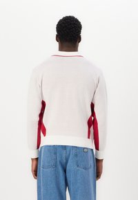 Stickad tröja i ljusgrått med röda sidosträck och en kontrasterande krage. Har ribbade muddar och midja, tillverkad av mjukt material.