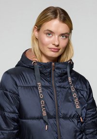 Marinblå pufferjacka med huva, med ett kviltat mönster, bronsfärgad dragkedja och tryckta snoddar med texten "POLO CLUB". Slät, glänsande textur.