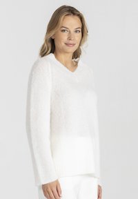 Kobieta z blond włosami do ramion, ubrana w biały, włochaty sweter w serek i białe spodnie, stoi na tle jasnego, jednolitego tła.