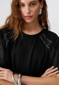 Črna majica s poudarjenim okroglim izrezom in satinastimi napihnjenimi rokavi. Opremljena s kovinskimi sponkami ob ramenih. Gladek tekstura.