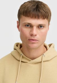 Sudadera beige hecha de tela suave, con capucha ajustable y ojales de metal plateado. El modelo tiene el cabello corto y una expresión neutral.