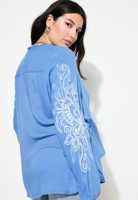 Blusa blu a maniche lunghe con ricami bianchi floreali sulle maniche e un collo a balze. Tessuto morbido con una vestibilità comoda.