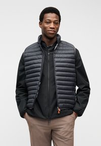 Άνδρας που φοράει μαύρο καπιτονέ γιλέκο πάνω από μαύρο μπουφάν με φερμουάρ και μπεζ παντελόνι, στέκεται με τα χέρια στις τσέπες μπροστά από λευκό φόντο.