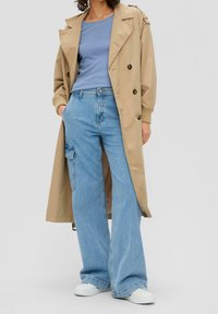 Beige trench coat dengan kancing ganda, atasan biru muda, celana jeans denim lebar warna cerah dengan saku samping, dan sepatu sneaker putih.