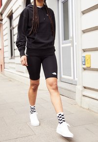 Osoba kráčející po chodníku, oblečená v černé mikině s kapucí, černých cyklistických kraťasech The North Face, bílých teniskách a černo-bílých kostkovaných ponožkách.