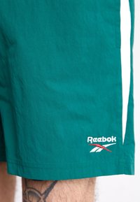 Teal šortky Reebok, vyrobené z lehkého materiálu, s bílým bočním pruhům a vyšitým logem blízko lemu. Texturovaný, casual design.