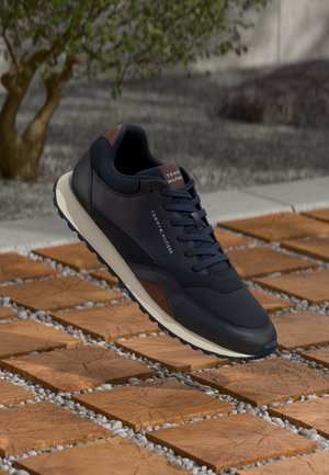 Navy blue sneaker with brown accents, mesh and leather materials, textured surface, and a white midsole, shown on wooden tiles with gravel.