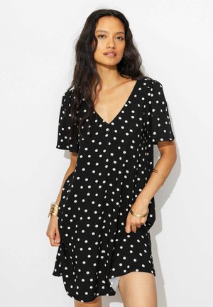 Femme aux longs cheveux foncés portant une robe noire à manches courtes avec des pois blancs et des bracelets dorés aux deux poignets.