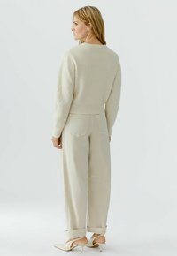 Vrouw staand in een beige gebreide trui, crèmekleurige broek met wijde pijpen en crèmekleurige slingback hakken, licht naar links gericht, met vooral haar achterkant zichtbaar.