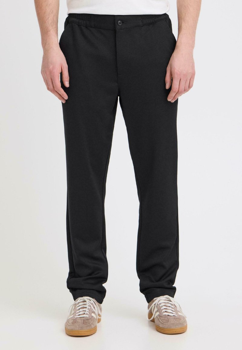 Homme portant un pantalon noir droit et des baskets beige, debout devant un fond blanc uni.