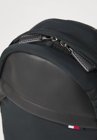 Svart ryggsekk med et glatt lærtopplag og stoffkropp. Har glidelås og en liten trikolor-logo nederst.