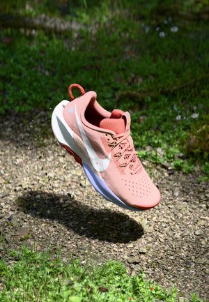 Light pink running shoe with a mesh upper, white swoosh logo, textured laces, and a rubber sole. Elevated above gravel and grass surface.
