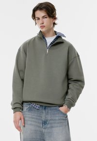 Felpa verde oliva con colletto a mezzo zip, design oversize e polsini a coste, indossata sopra una camicia a quadri blu, abbinata a jeans in denim chiaro.