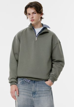 Sweatshirt vert olive avec un col zippé demi-long, design oversize et poignets côtelés, superposé sur une chemise à carreaux bleus, porté avec un jean en denim clair.