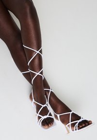 Sandali con tacco alto bianchi e con cinturino, caratterizzati da sottili lacci che si incrociano sulle gambe, con una punta quadrata e un delicato tacco a stiletto.