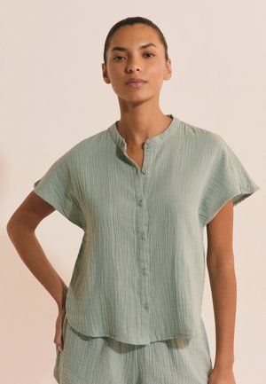 Kobieta stoi z jedną ręką na biodrze, ubrana w jasnobeżową krótki rękaw koszulę zapinaną na guziki i pasujące szorty na neutralnym tle.