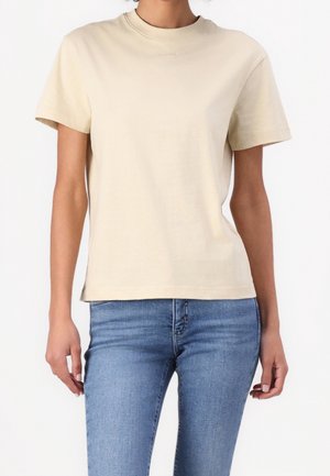 Person trägt ein schlichtes beiges Kurzarm-T-Shirt und blaue Jeans und steht vor einem weißen Hintergrund.