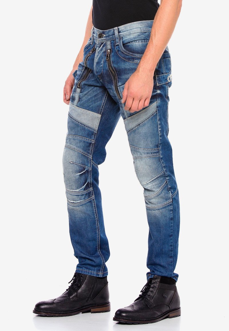 Blaue Jeans aus Denim mit einem Paneel-Design, sichtbaren Nähten, abgenutzten Akzenten und mehreren Reißverschluss-Taschen, kombiniert mit schwarzen Stiefeln.