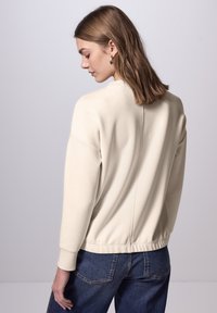 Felpa color crema con una vestibilità rilassata, polsini e vita a costine, caratterizzata da una cucitura centrale sul retro. Abbinata a jeans di denim.