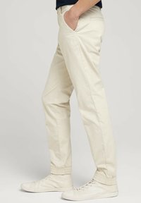 Jasnobeżowe chinosy z elastycznymi mankietami, przednimi kieszeniami i dopasowanym krojem. Połączone z białymi, wysokimi sneakersami.