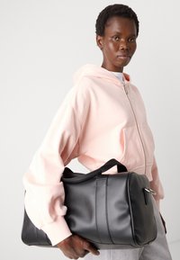 Borsa duffel nera con texture in pelle liscia, forma tondeggiante, maniglie doppie e dettagli in metallo argento. Il modello indossa un hoodie rosa chiaro con zip.