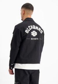 Zwarte denimjas met geborduurde witte letters op de achterkant, met "BLCKBNNS" en "BAD HABITS." Inclusief knoopmanchetten en een verkorte stijl.