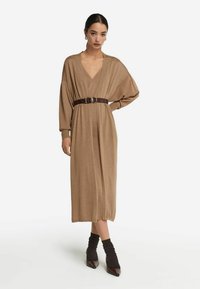 Brun långärmad klänning med djup V-ringning, rynkad tyg och ett brunt bälte. Har en lös passform och når under knäna.