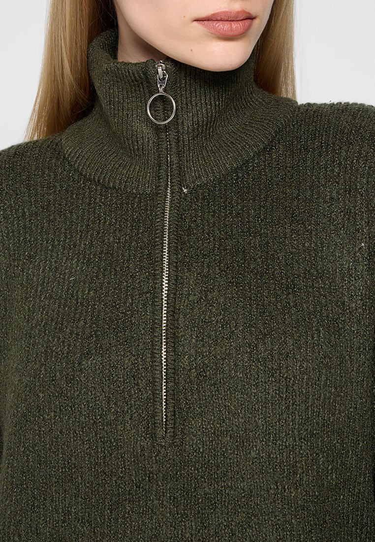 Olivgrüner Strickpullover mit hohem Kragen und Reißverschluss, aus strukturiertem Stoff mit silberfarbener Reißverschlussbeschlägen.