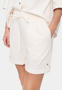 Shorts en lin blancs avec ceinture à cordon de serrage, manches retroussées et accents de boutons dorés. Texture lisse et coupe ample pour le confort.