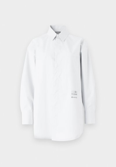 Valge pikakä sleeve shirt, mis on valmistatud kerge kangast; omab nööpidega krae, lõdva istuvuse ja trükitud logo detaili külgedel.