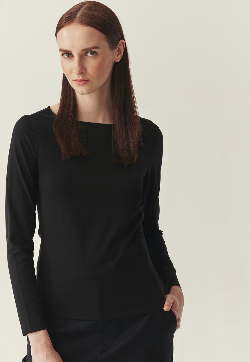Haut noir à manches longues avec un décolleté carré, une texture lisse et une coupe ajustée. Le modèle pose avec une main dans la poche d'un pantalon marine.