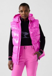 Gilet imbottito rosa con finitura lucida e cappuccio, abbinato a un maglione nero e pantaloni rosa. Presenta tasche con zip e cuciture visibili.