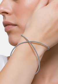 Bracciale in metallo argentato con un design liscio e flessibile, caratterizzato da bande incrociate che creano un aspetto minimalista.