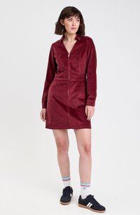 Robe en velours côtelé rouge avec des manches longues, fermeture éclair à l'avant, col pointu et taille cintrée, associée à des baskets sombres et des chaussettes rayées.