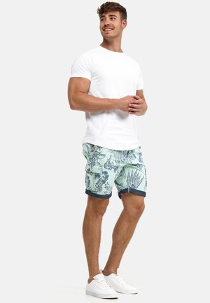 Young man standing with hands clasped, wearing a white t-shirt, light blue floral cargo shorts, and white sneakers.