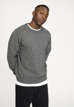Grå strikket sweater med rund halsudskæring og ribbede manchetter, lagdelt over en hvid T-shirt, båret med sorte bukser. Glat tekstur.