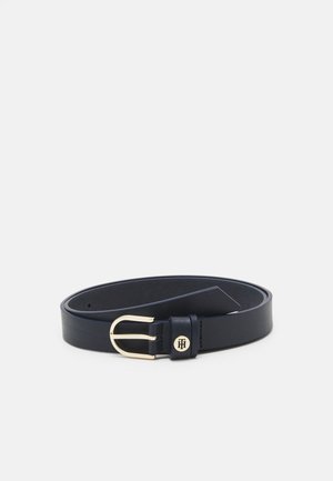 Ceinture en cuir noir avec boucle arrondie dorée et petit détail de logo circulaire sur la boucle de la lanière, présentée enroulée sur fond blanc.