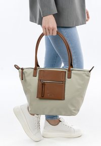 Beige tote bag with brown leather accents, featuring a front zip pocket and two top handles. Material appears smooth and durable.