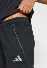 Pantaloni sport negri cu finisaj texturat, având o dungi laterală gri, buzunar frontal și logo-ul Adidas alb pe partea inferioară a piciorului.