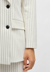 Valkoinen pinstripe-puku, jossa mustat napit, joka on muotoiltu istuvaksi, notched-lapeli ja yksi sivutasku, joka esittelee sileää kankaan rakennetta.