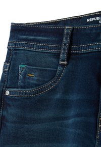 Vaqueros de mezclilla azul oscuro con un pequeño bolsillo frontal, costura dorada y una etiqueta de marca visible en la cintura. Textura suave, diseño clásico.