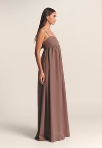 Bruine, vloerlange jurk met dunne bandjes en een getailleerd lijfje. Materiaal lijkt glad met een lichte glans en plooit bij de taille.