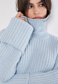 Maglione a coste di un blu chiaro con un collo alto e maniche lunghe e oversize. Presenta una trama a maglia spessa e dettagli di polsini larghi.