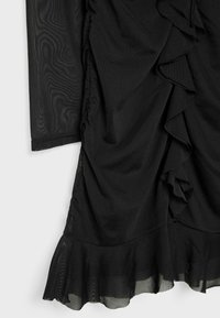 Černé šaty s vrstvenými volány, hladkou texturou a průsvitným detailem na rukávech. Spodní lem má jemný volán. Tkanina vypadá lehce a splývavě.