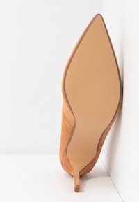 Chaussure à talon haut en daim marron clair à bout pointu, positionnée semelle vers le haut contre un mur et un sol blancs en angle.