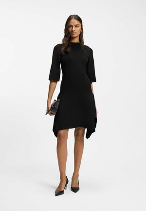 Kvinde med langt mørkt hår iført en sort knælængde kjole med trekvartærmer og sorte hæle, der holder en lille sort håndtaske.